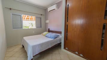 Quarto com cama, guarda-roupa de madeira, ar condicionado e janela com persianas.