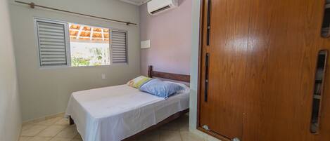 A bedroom with a bed, a wooden wardrobe, an air conditioning unit, and a window with shutters.