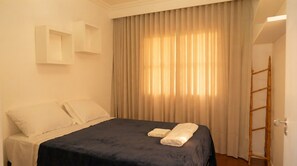 Hotellihuone, jossa on sänky, ikkuna verhoilla, ovi ja seinään kiinnitetty valaisin.