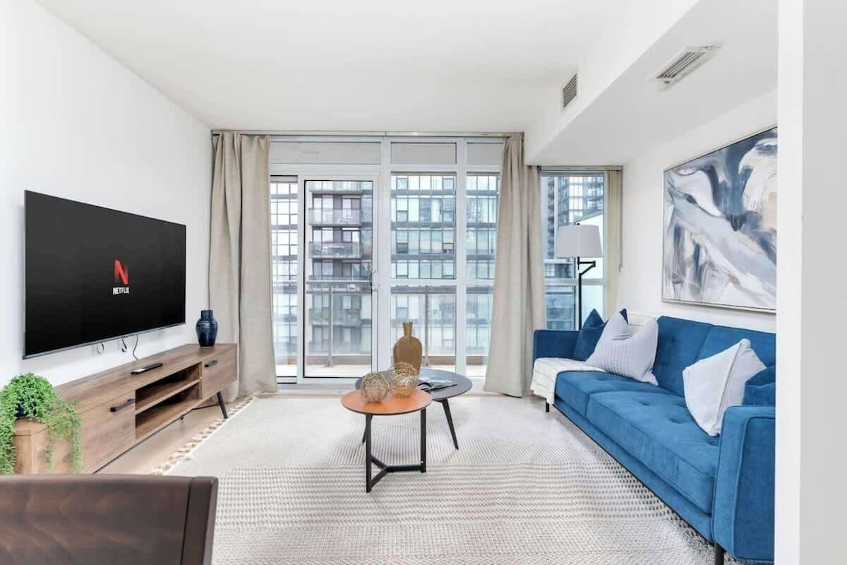 A modern living room with a flat-screen TV, a blue sofa, a coffee table, and a large window with a view of buildings.