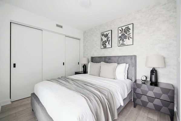 Une chambre à coucher comprenant un lit, deux tables de chevet, une commode, une armoire et deux tableaux représentant des fleurs, accrochés au mur.