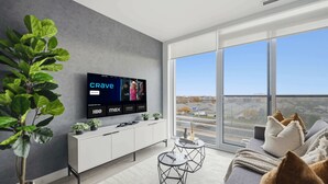 A modern living room with a large flat-screen TV displaying a streaming service interface, a white entertainment unit, a round coffee table, and a large window offering a city view.