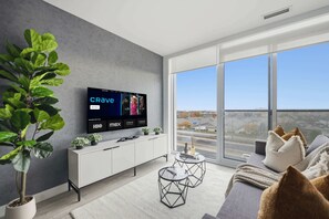 Un salon moderne doté d’un grand téléviseur à écran plat affichant l’interface d’un service de diffusion en continu, d’un meuble de rangement blanc, d’une table basse ronde et d’une grande fenêtre offrant une vue sur la ville.