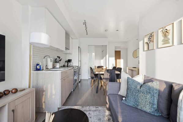 A modern kitchen with white cabinets, a dining area with a table and chairs, and a living room with a sofa and cushions.