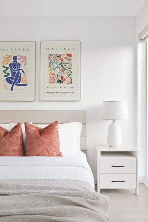 A bedroom with a bed, pillows, a nightstand, a lamp, and two framed artworks on the wall.