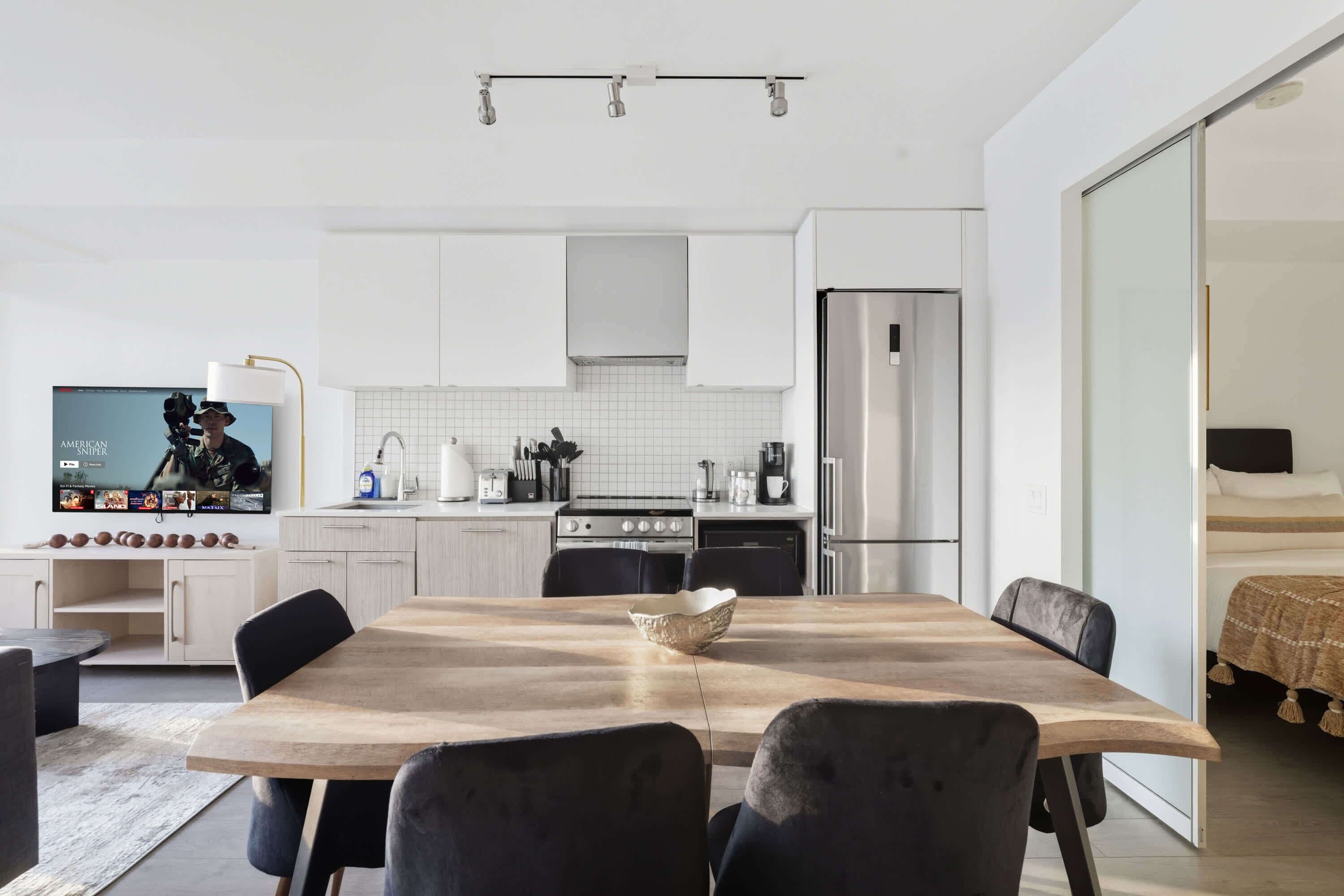 A modern kitchen with white cabinets, a stainless steel refrigerator, and a dining area with a wooden table and black chairs.