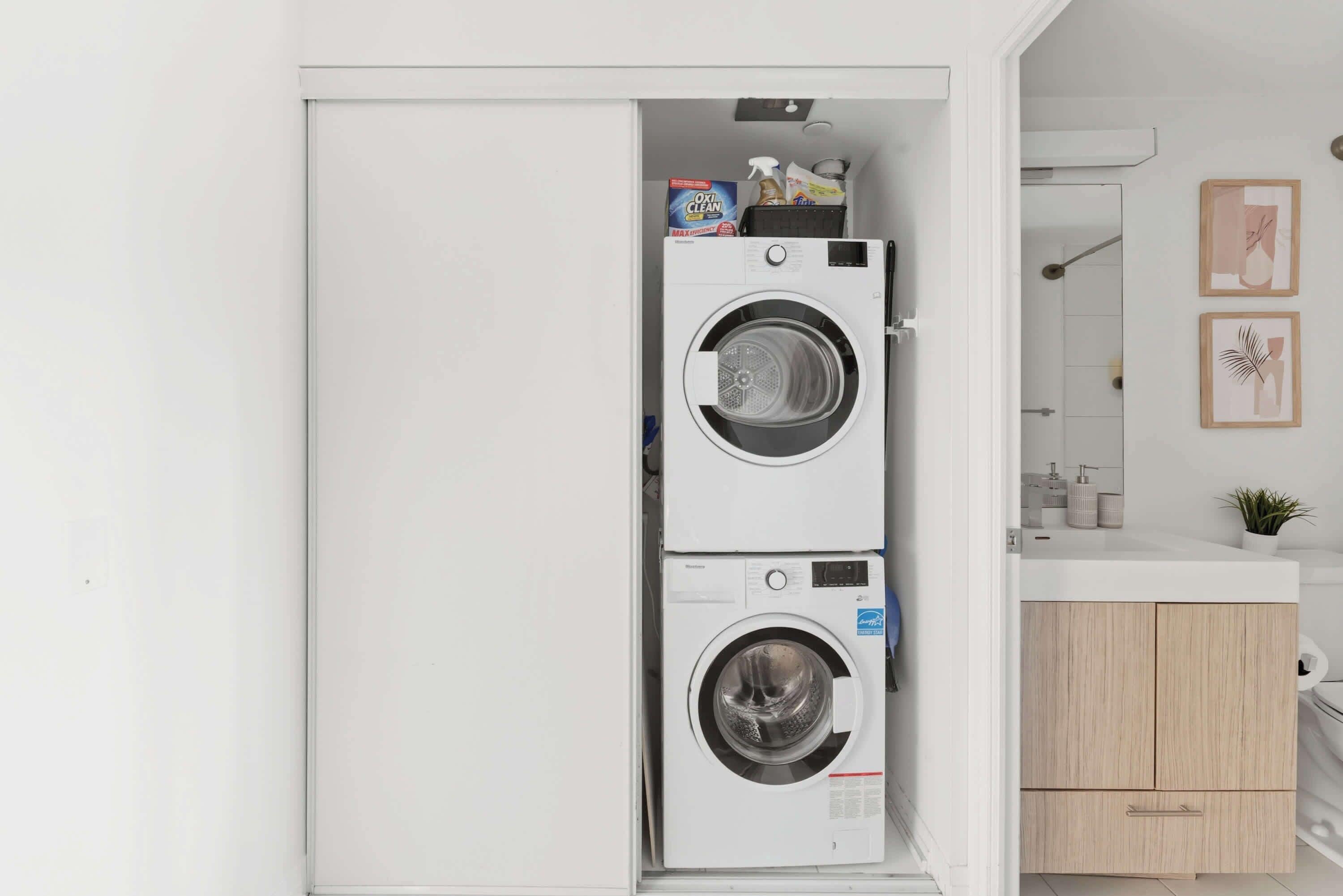 A compact laundry room with two stacked front-loading washing machines, a small sink, and a wooden cabinet.