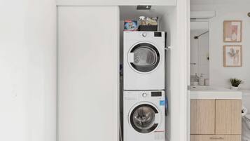 A compact laundry room with two stacked front-loading washing machines, a small sink, and a wooden cabinet.