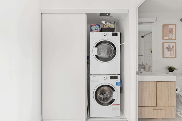 A compact laundry room with two stacked front-loading washing machines, a small sink, and a wooden cabinet.