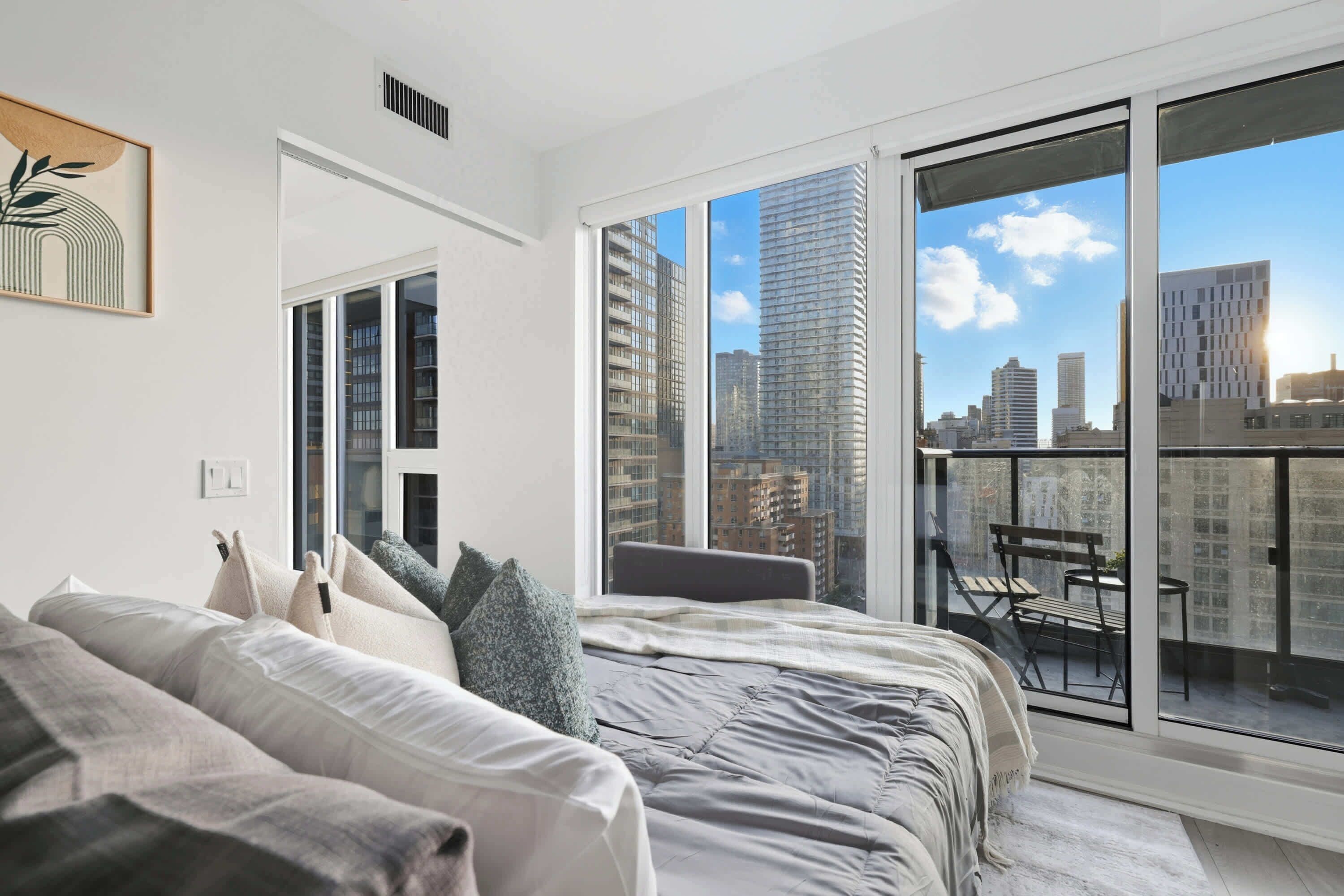 A modern bedroom with a large bed, a view of the cityscape through floor-to-ceiling windows, and a balcony with a table and chairs.