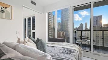 A modern bedroom with a large bed, a view of the cityscape through floor-to-ceiling windows, and a balcony with a table and chairs.