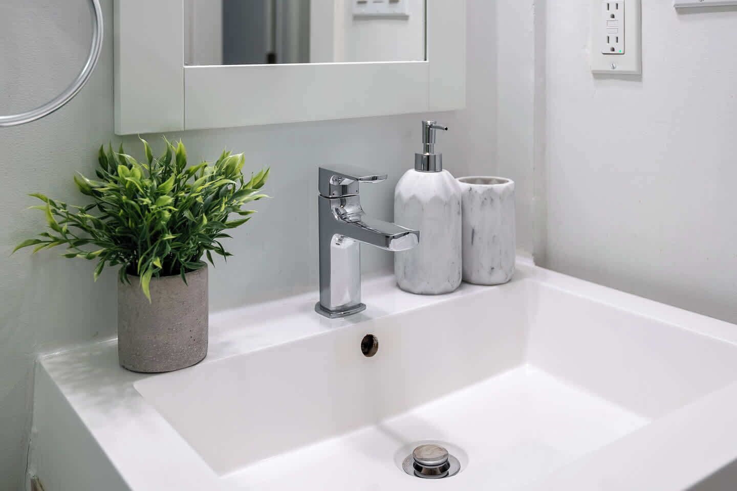 A bathroom with a white sink, a chrome faucet, a potted plant, and two marble containers.