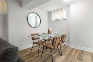 Ein moderner Essbereich mit einem Glastisch, braunen Polsterstühlen, einem runden Spiegel an der Wand und einem Fenster, das für natürliches Licht sorgt.