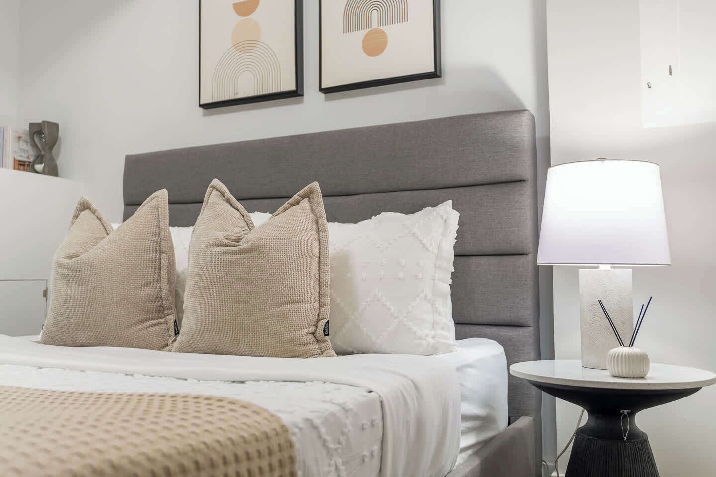 A neatly made bed with a grey headboard, a bedside table with a lamp, and two framed artworks on the wall.