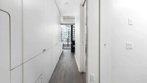 Un couloir étroit aux murs blancs et au plancher en bois, qui mène à une pièce dotée d’une grande fenêtre.