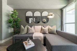 Ein modernes Wohnzimmer mit einem grauen Ecksofa, einem schwarzen Couchtisch mit Zeitschrift und Gläsern, einer Stehlampe und einem großen Spiegel mit drei Teilen.
