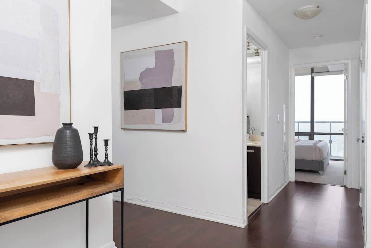 A modern interior with a hallway leading to a bedroom. There is a console table with a vase and candle holders. A framed abstract painting is on the wall.