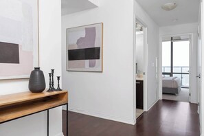 Ein modernes Interieur mit einem Flur, der zu einem Schlafzimmer führt. Eine Konsole mit einer Vase und Kerzenständern steht dort. Ein gerahmtes, abstraktes Gemälde hängt an der Wand.