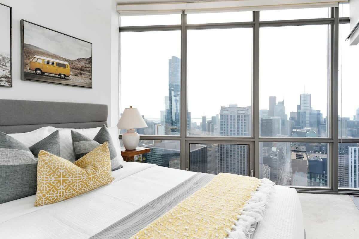Una camera da letto con un'ampia finestra, un letto con cuscini decorati, una lampada da comodino e vista sulla città.