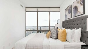 A modern bedroom with a large bed, a tufted headboard, and a city view through floor-to-ceiling windows. - Above the City Designer Sub-Penthouse Oasis (Toronto)