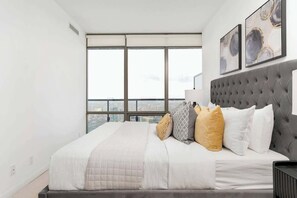 Ein modernes Schlafzimmer mit einem großen Bett, einer gesteppten Kopfteil und Blick auf die Stadt durch raumhohe Fenster.