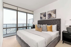 A bedroom with a large bed, a nightstand, a lamp, and a view of the city through large windows.