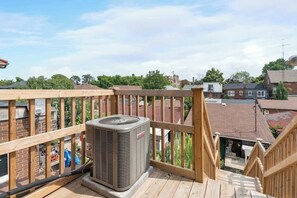 A wooden deck with a view of a residential area, a central air conditioning unit, and a staircase leading to an upper level.