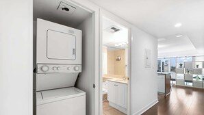 Une buanderie moderne équipée d’un lave-linge et d’un sèche-linge à chargement frontal, une salle de bain avec des toilettes et un lavabo, et une vue sur le paysage urbain grâce à de grandes fenêtres.