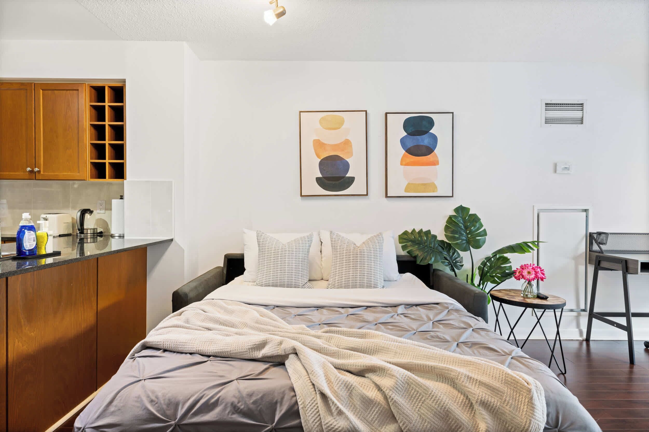 A modern bedroom with a bed, a small side table, a potted plant, and artwork on the wall.