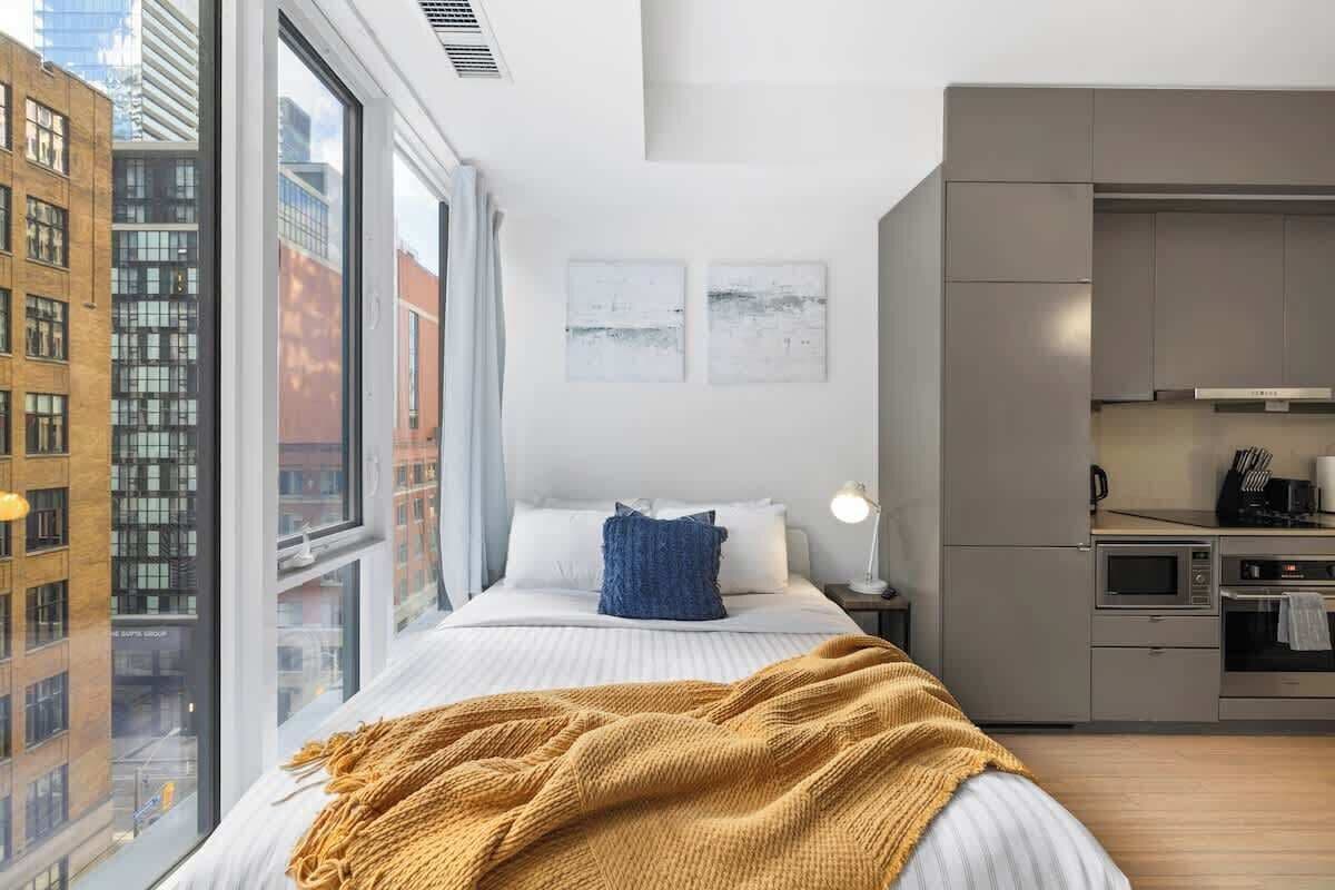 Une chambre moderne dotée d’une grande fenêtre, d’un lit recouvert d’une couverture jaune, d’une table de chevet avec une lampe et d’une kitchenette équipée d’appareils électroménagers.