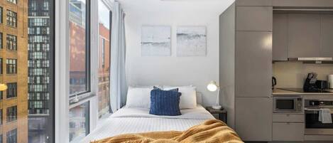 Une chambre moderne dotée d’une grande fenêtre, d’un lit recouvert d’une couverture jaune, d’une table de chevet avec une lampe et d’une kitchenette équipée d’appareils électroménagers.