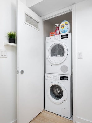 A compact laundry room with two stacked washing machines, a shelf with a plant, and a large bottle of Tide detergent. - Cozy Toronto Stay Skyline View | Parking Available (Toronto)