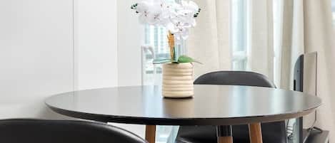 A round dining table with a vase of white flowers, black chairs, and a view of a cityscape through large windows.
