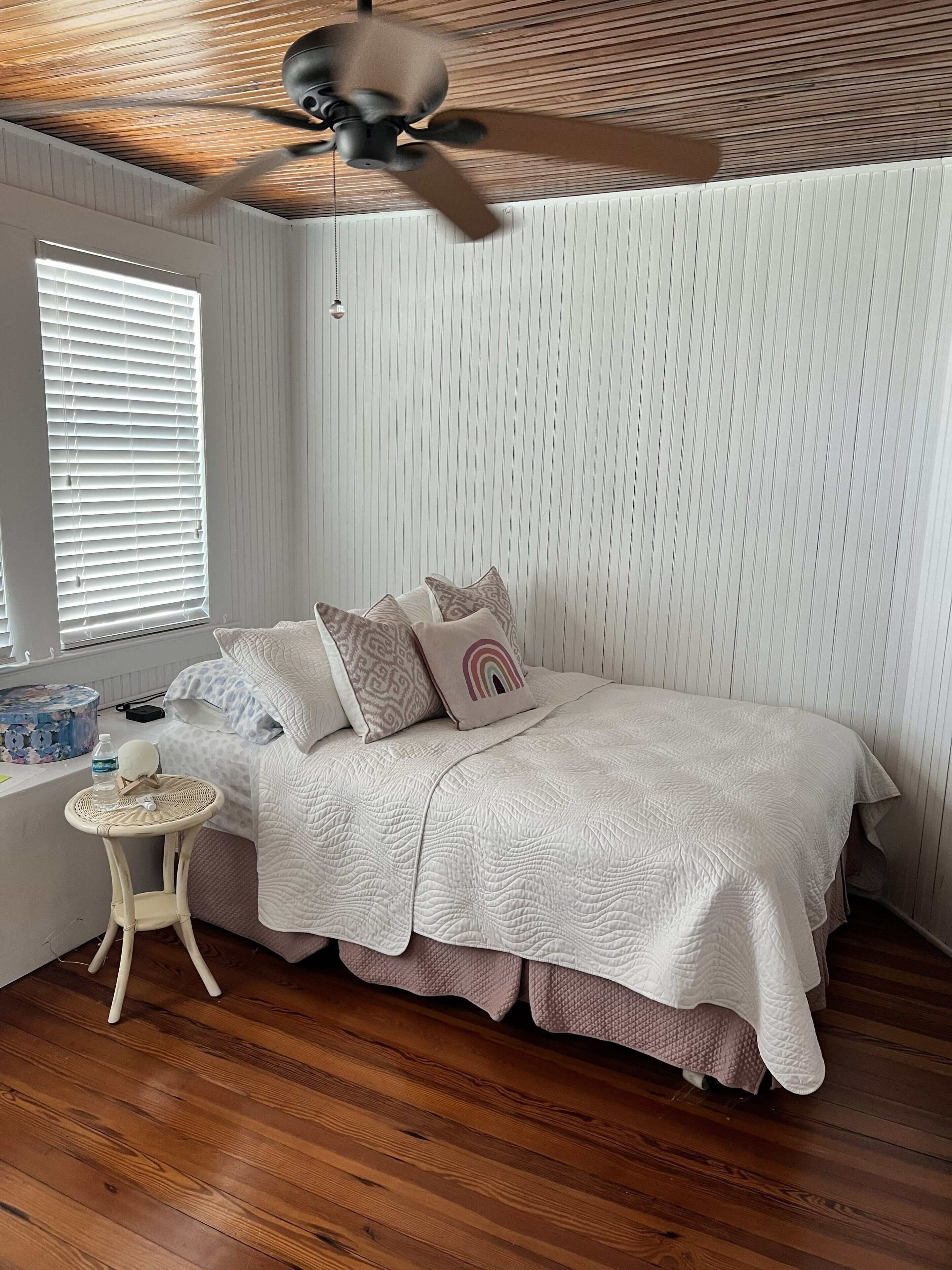 Une chambre à coucher avec un lit, des oreillers, une petite table, un ventilateur de plafond et une fenêtre avec des stores.