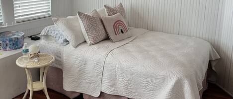 Un dormitorio con cama, almohadas, una mesita, ventilador de techo y una ventana con persianas.