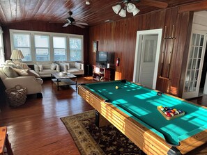 A pool table with balls, a sofa, a television, and a ceiling fan in a room with wooden walls and windows. - Historic waterfront 4-bedroom, 2 bath house on St Lucie River-Big Water Views (Stuart)