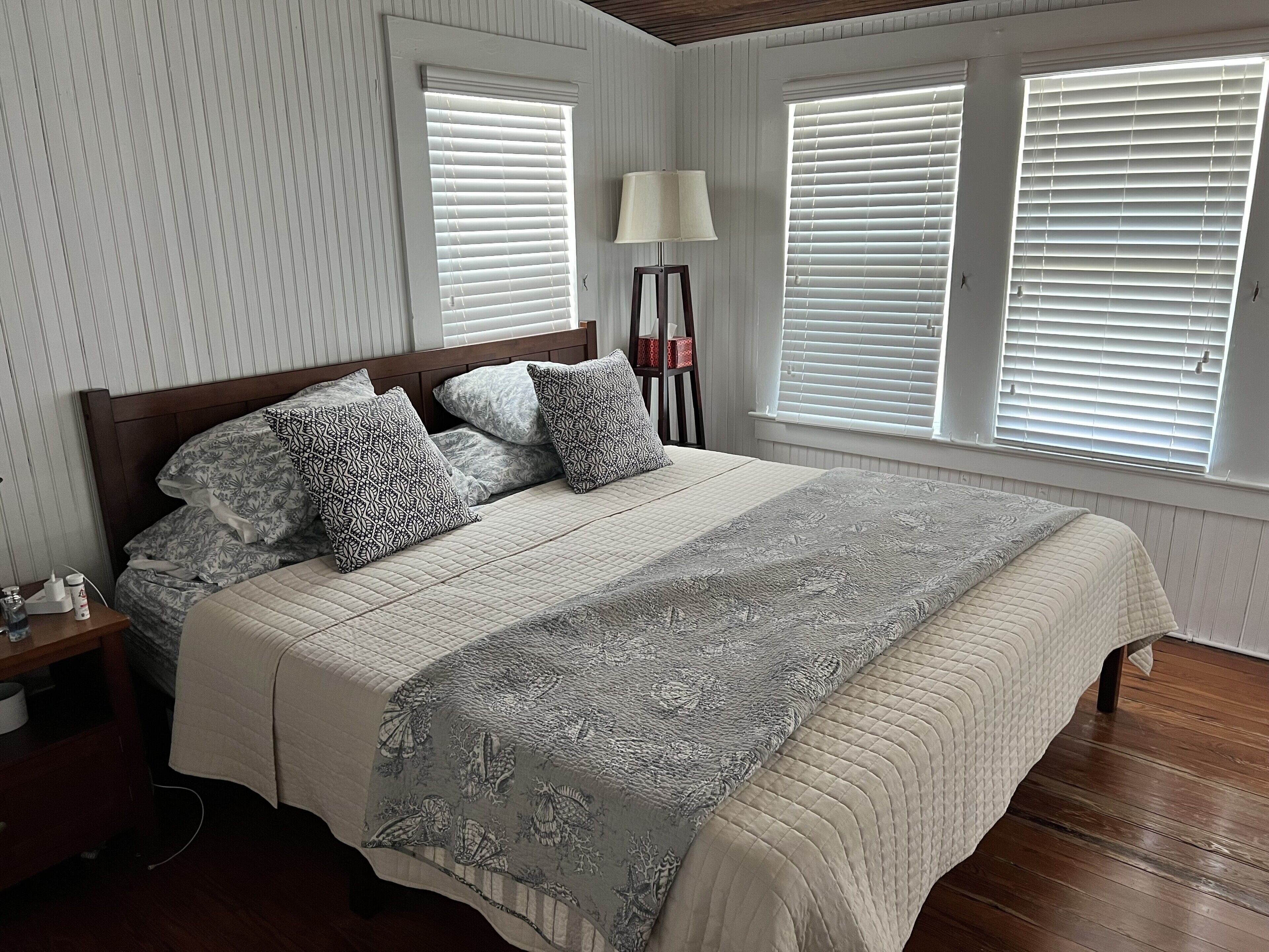 Ein Schlafzimmer mit Bett, Nachttisch, Lampe und Fenstern mit Jalousien.