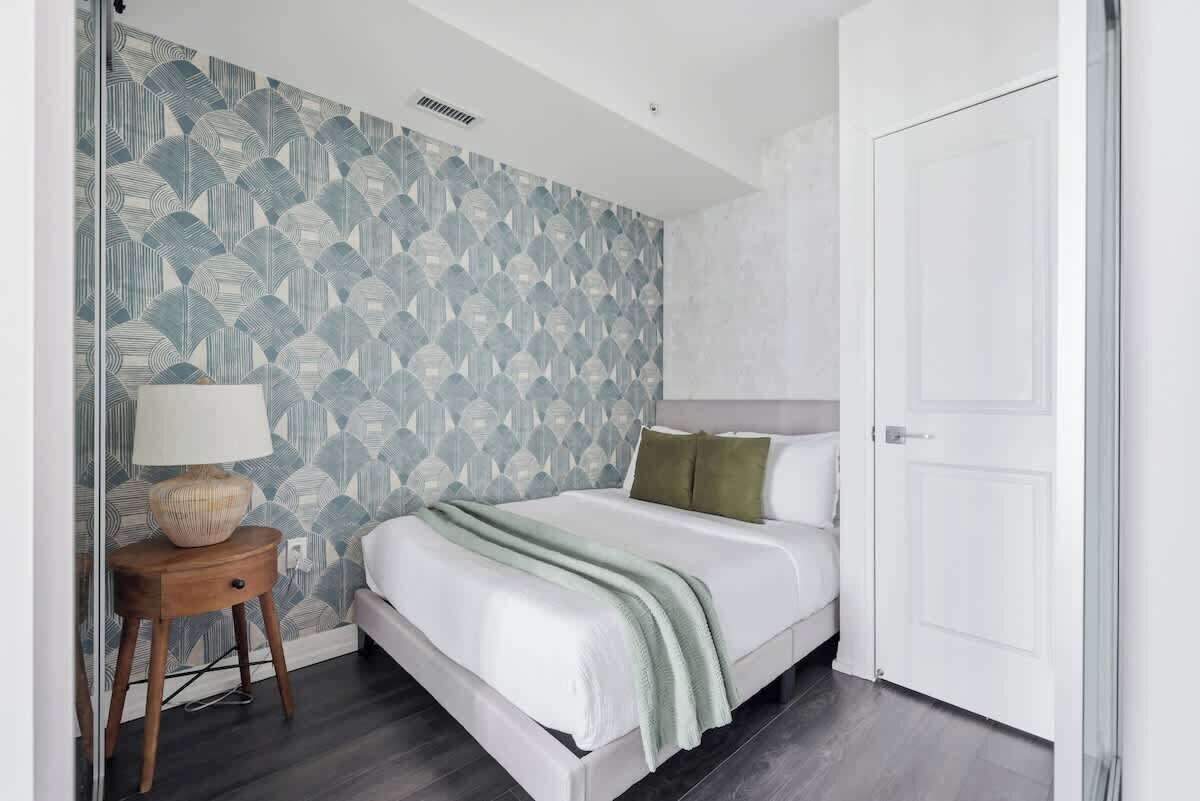A bedroom with a bed, a bedside table, a lamp, and a patterned wallpaper.