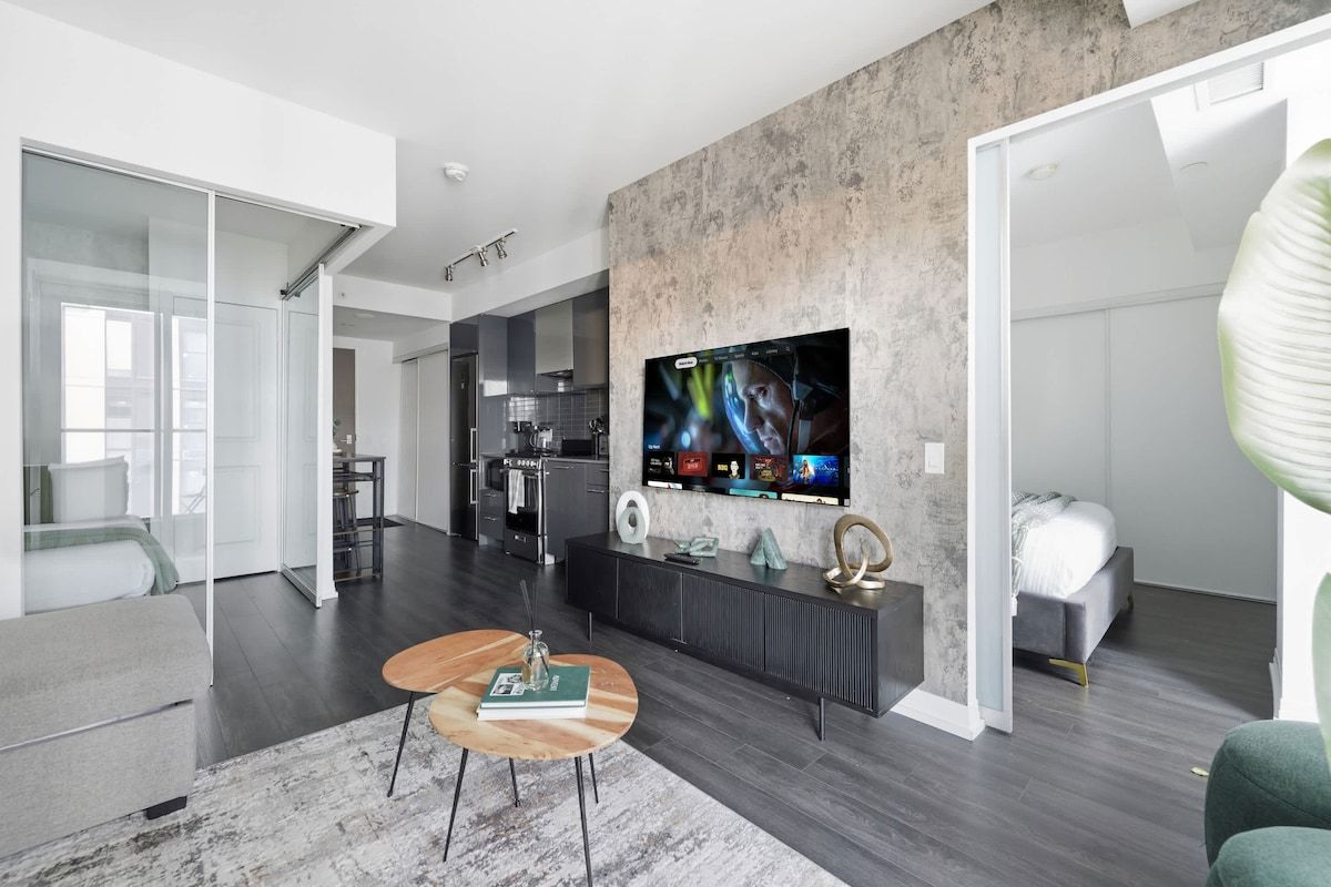 A modern living room with a large flat-screen TV mounted on a textured wall, a sofa, a coffee table, and a kitchen in the background.