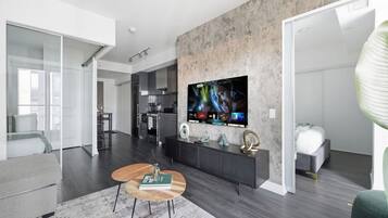 A modern living room with a large flat-screen TV mounted on a textured wall, a sofa, a coffee table, and a kitchen in the background.