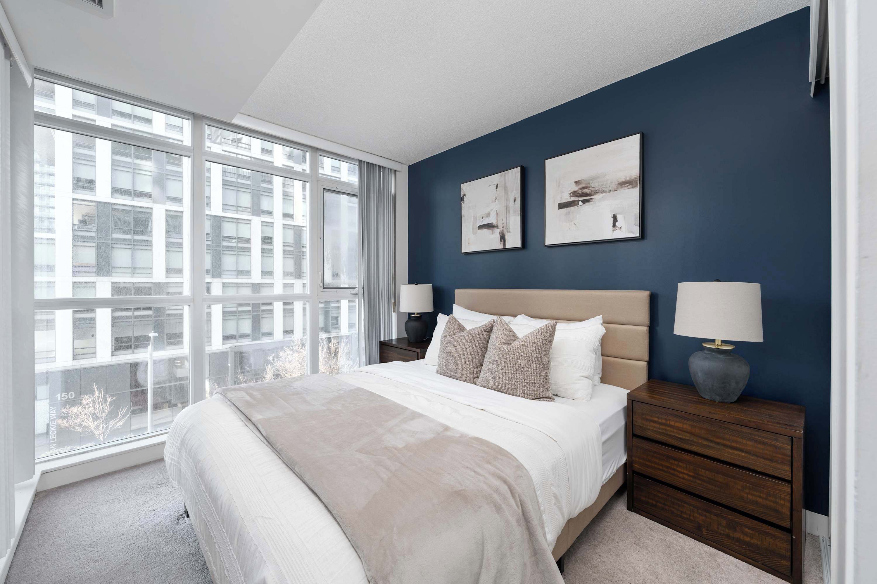 A bedroom with a large window, a bed with a beige headboard, two bedside tables with lamps, and two wall-mounted abstract paintings.