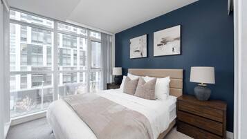 A bedroom with a large window, a bed with a beige headboard, two bedside tables with lamps, and two wall-mounted abstract paintings.