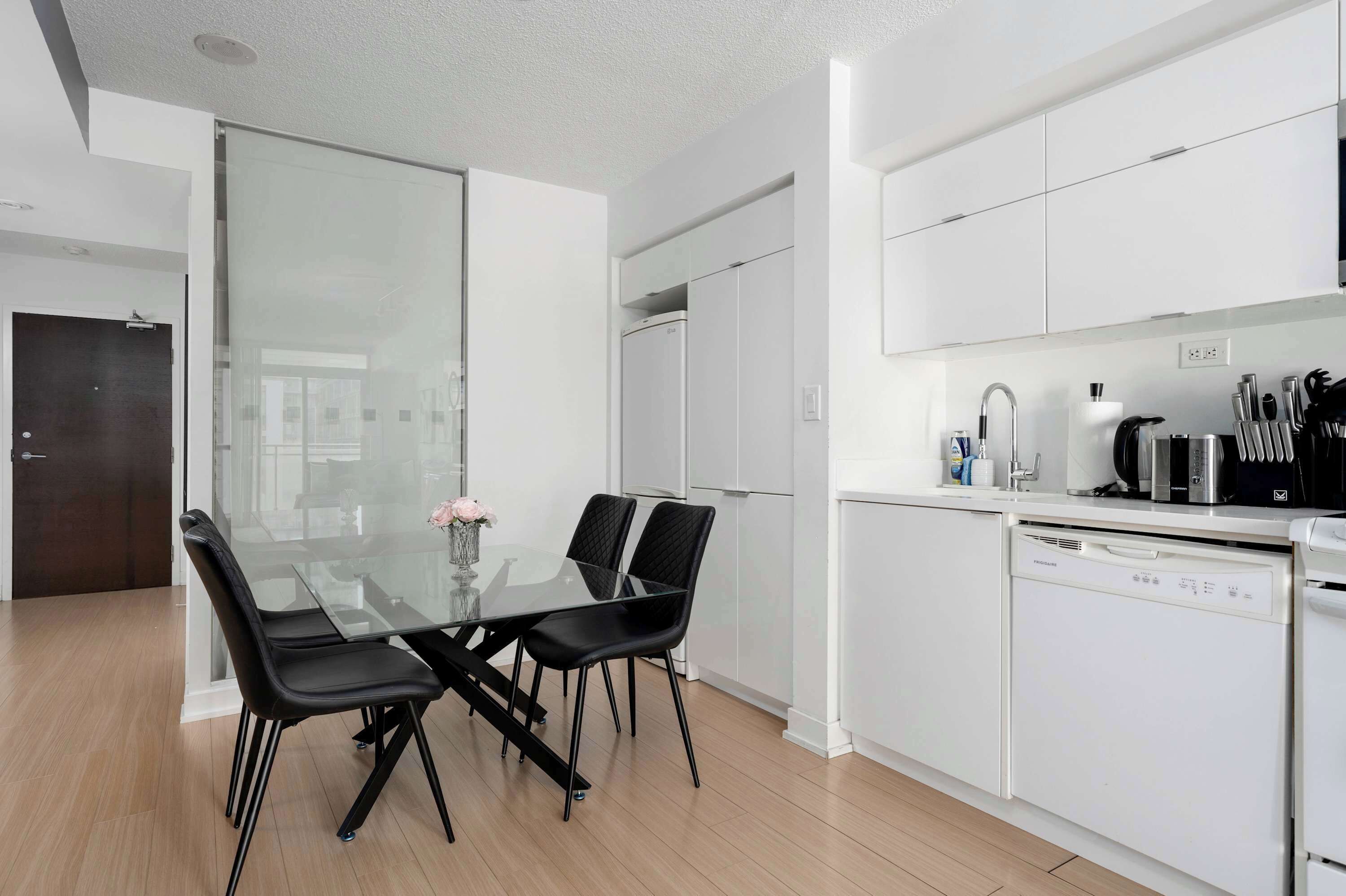 A modern kitchen with white cabinetry, a glass dining table with black chairs, and a dishwasher.