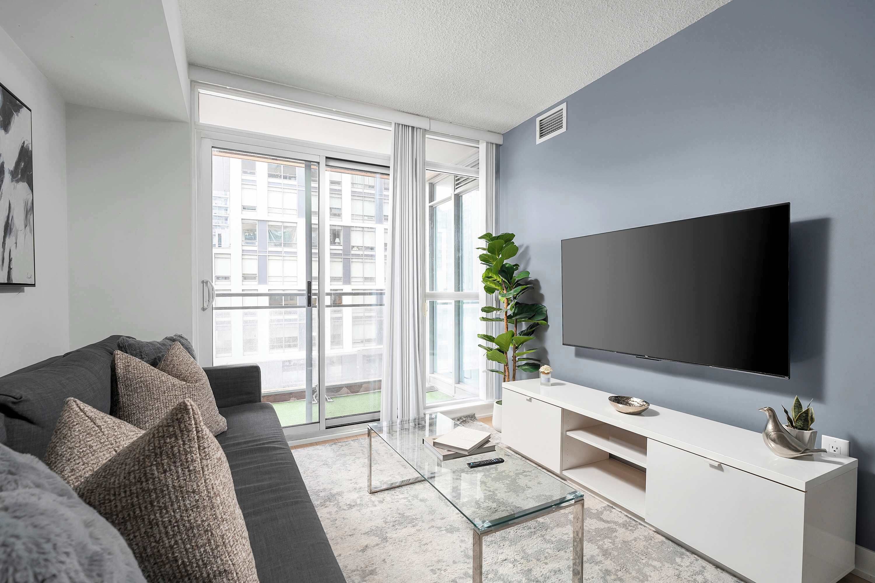 Un salon moderne avec un canapé gris, une table basse en verre, un meuble de rangement blanc pour appareils électroniques et un grand téléviseur à écran plat.