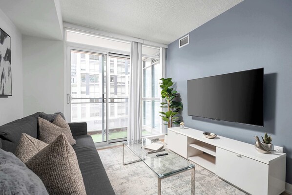 Un salon moderne avec un canapé gris, une table basse en verre, un meuble de rangement blanc pour appareils électroniques et un grand téléviseur à écran plat.