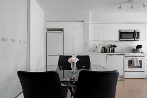A modern kitchen with white cabinetry, a microwave, a coffee maker, and a dishwasher. There is a glass dining table with two chairs and a vase with flowers. - Walk to Rogers Centre, CN Tower, Waterfront!  | Parking Available (Toronto)
