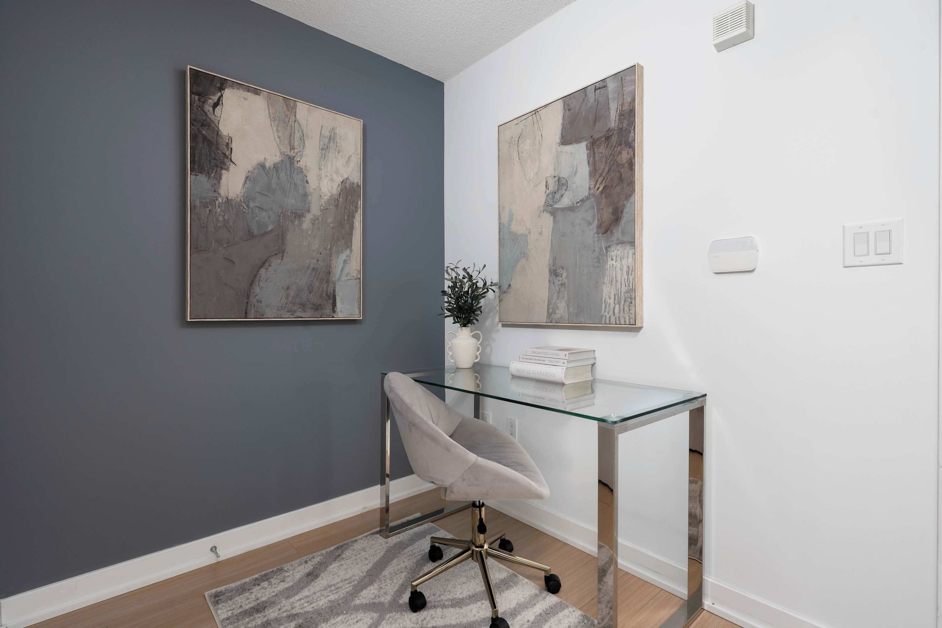 Een kamer met een glazen bureau, een grijze stoel en abstracte schilderijen aan de wanden.