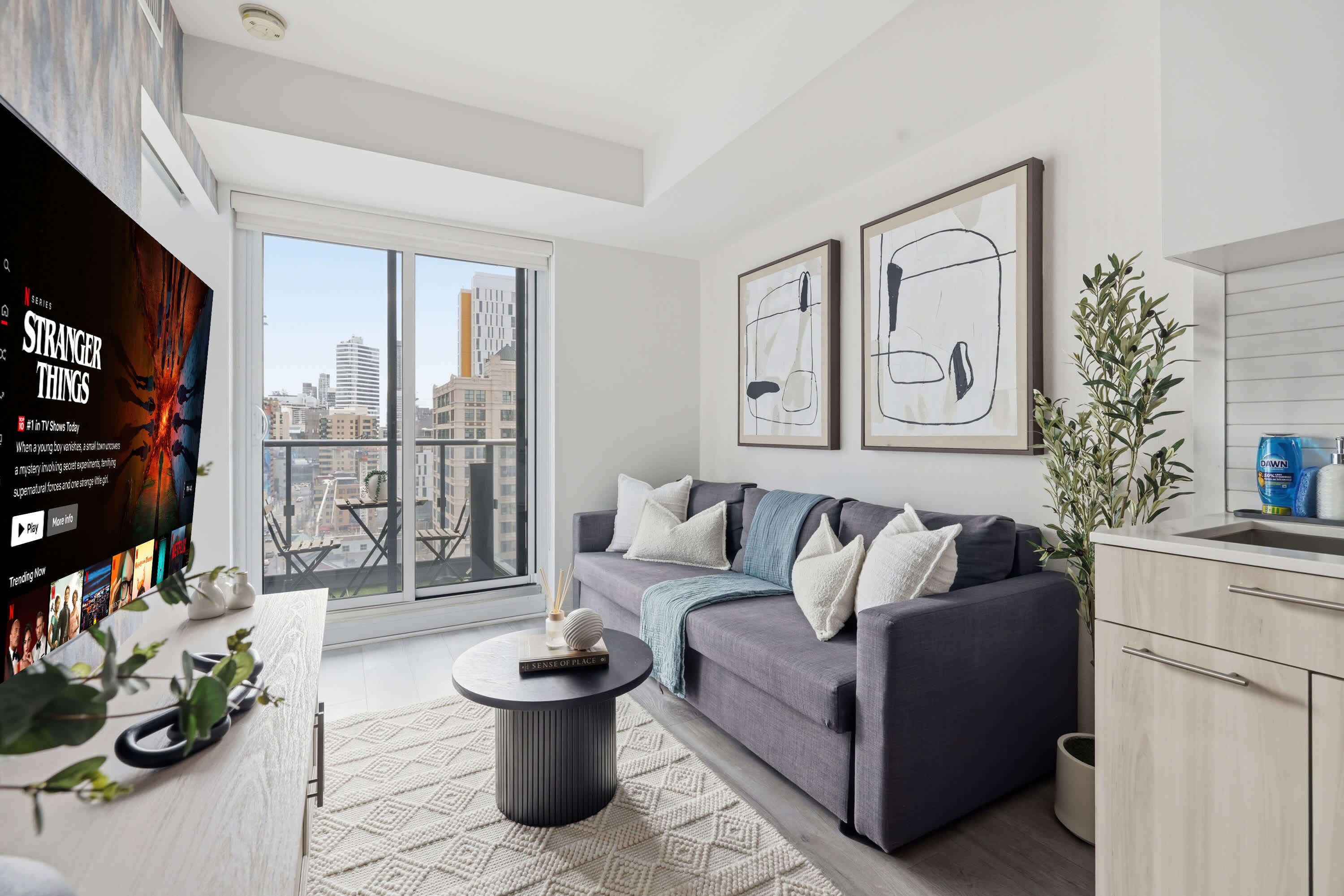 A modern living room with a grey sofa, a coffee table, and a large window with a city view.