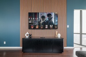 A modern living room with a large flat-screen TV mounted on a wooden wall, a dark wood console table in front, and a large window providing a view of the city. - City Lights & Skyline Nights, Bay Street Penthouse (Toronto)