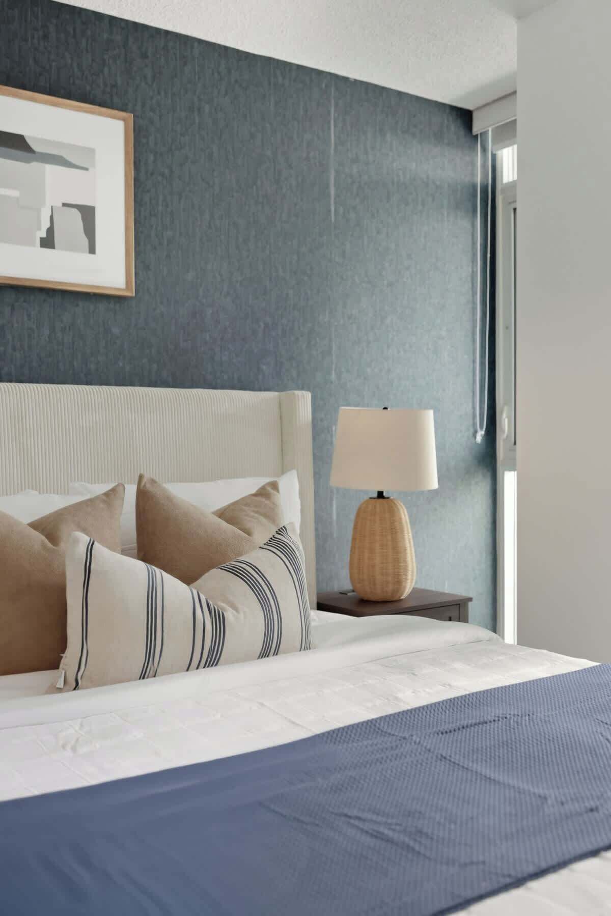 A neatly made bed with a blue bedspread and striped pillows, a bedside lamp on a wooden nightstand, and a framed picture on the wall.